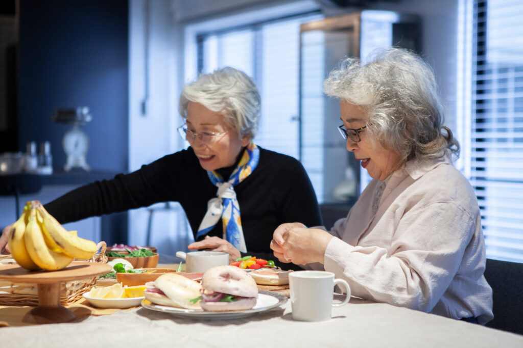 高齢の女性がビュッフェを楽しんでいる。テーブルにはバナナ、ハンバーガー、コーヒー、サンドイッチなどが並んでいる。